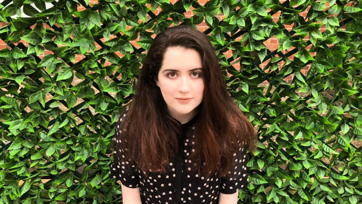 Head and shoulder portrait of a young woman with long brown hair standing in front of a wall with ivy plant