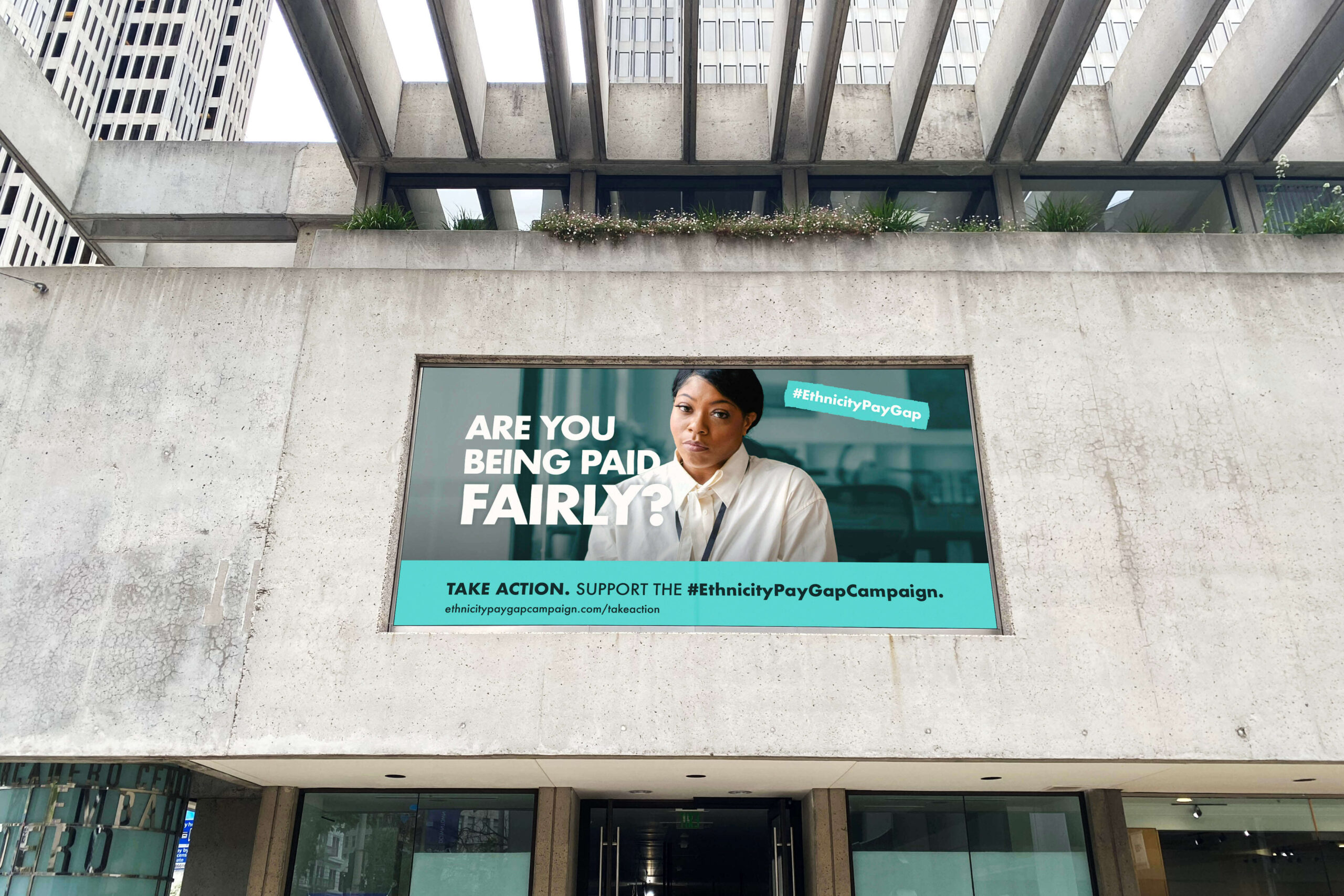 A large outdoor poster on a concrete building, featuring a campaign for pay equity. The poster shows a woman with a serious expression, accompanied by bold text reading 'Are you being paid fairly?' followed by '#EthnicityPayGap' in a hashtag label. Below, the message 'Take action. Support the #EthnicityPayGapCampaign.' directs viewers to the campaign's website link for more information. The poster uses a combination of teal, white, and black for a strong visual contrast.