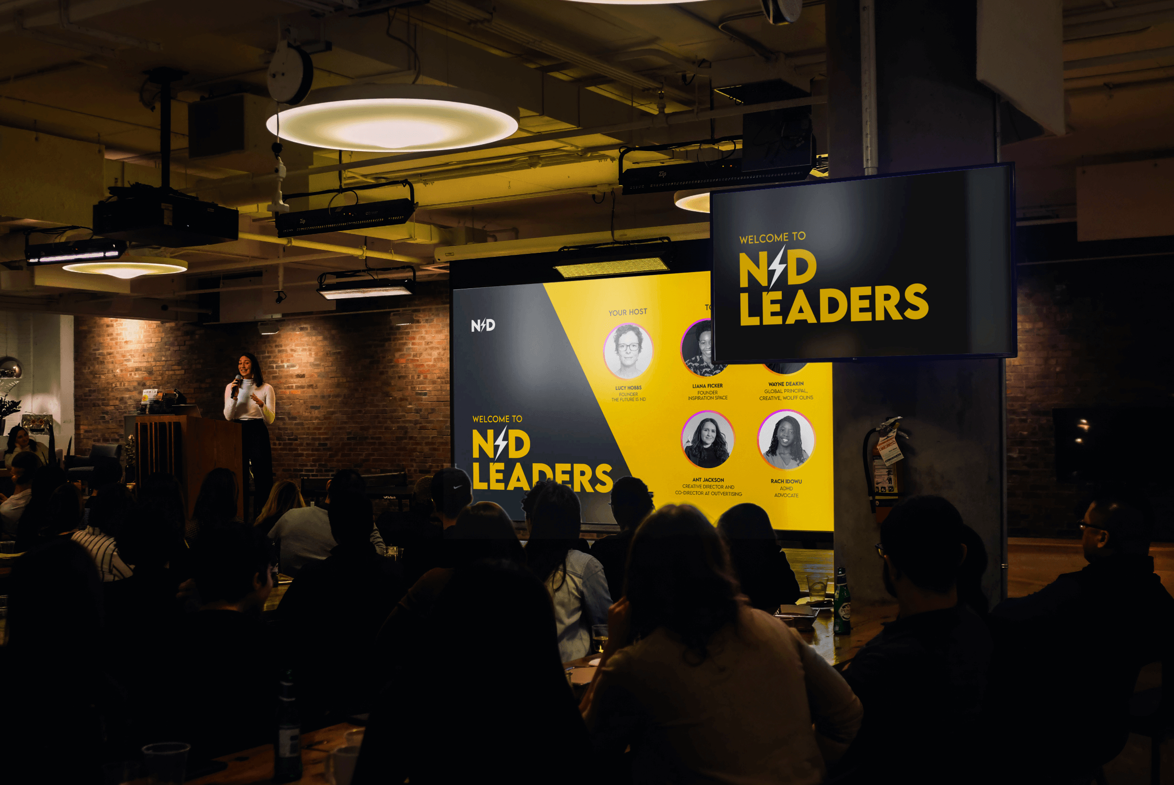 ND Leaders event in a modern, dimly lit venue. A large screen displays 'Welcome to ND Leaders' with host and speaker details. Attendees sit attentively, engaging in a discussion about neurodiversity in leadership.