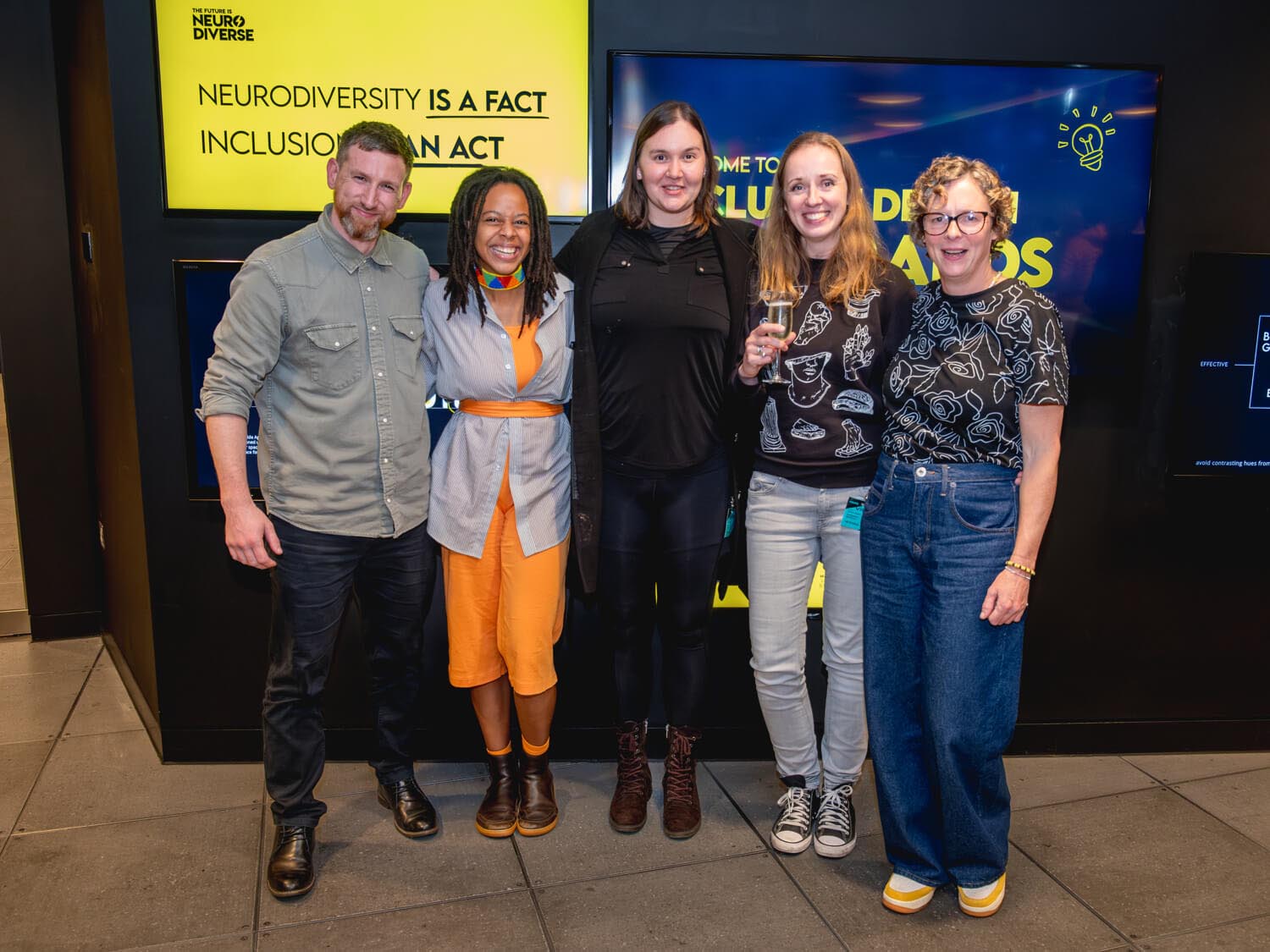 A group of five panelists smiling and standing together in front of a bright yellow and blue event backdrop. The backdrop displays the phrases "Neurodiversity is a fact" and "Inclusion is an act," alongside "Inclusive Design for Brands." The individuals are dressed casually, radiating a warm and inclusive atmosphere.