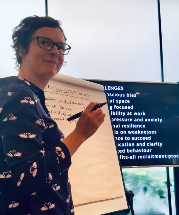 a shorthaired woman wearing glasses standing by a white board holding a pen.