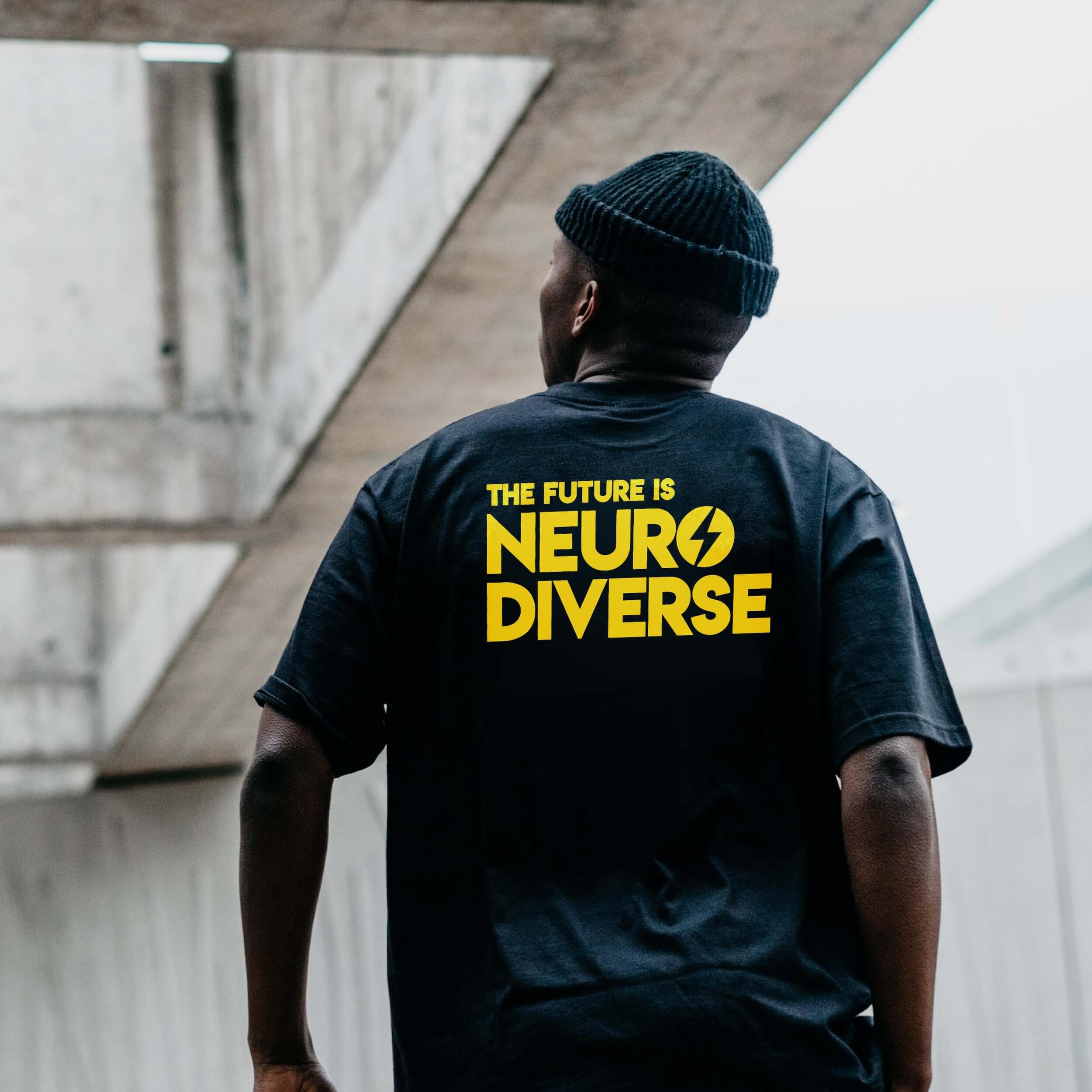 Person wearing a black T-shirt with 'The Future is Neurodiverse' in bold yellow text on the back, featuring a lightning bolt icon. The minimalist urban background reinforces a modern and inclusive brand identity.